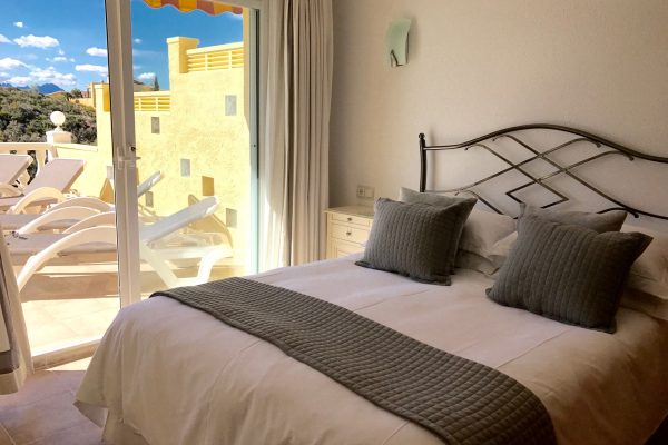 The king sized bed in the silver bedroom with decorative cushions and bed runner, sliding glass doors to the terrace with four sun loungers
